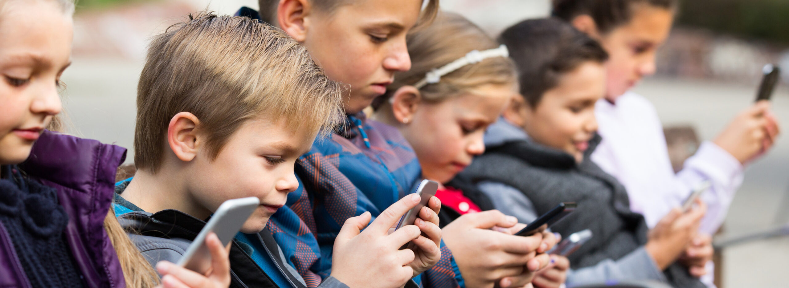 Six different kids standing in a line side by side, all looking down at their phones